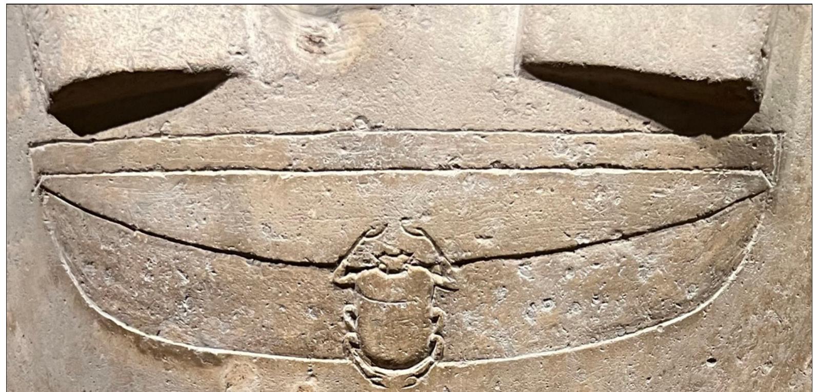 ndasma he chin is roken off  L, slightly smiling mouth. The large ears are carefully carved, anc rounded and adorned with a short divine beard (the end of which i:  with no internal markings. The edge of a garment is visible partway  lown the neck; below this, the body is shown as completely swathed with nc  rms visib nd both s  e. The joined legs are gently modeled, with the knees in higher relief hins can be clearly distinguished. The coffin sits on an integral plinth  urved in  front. 