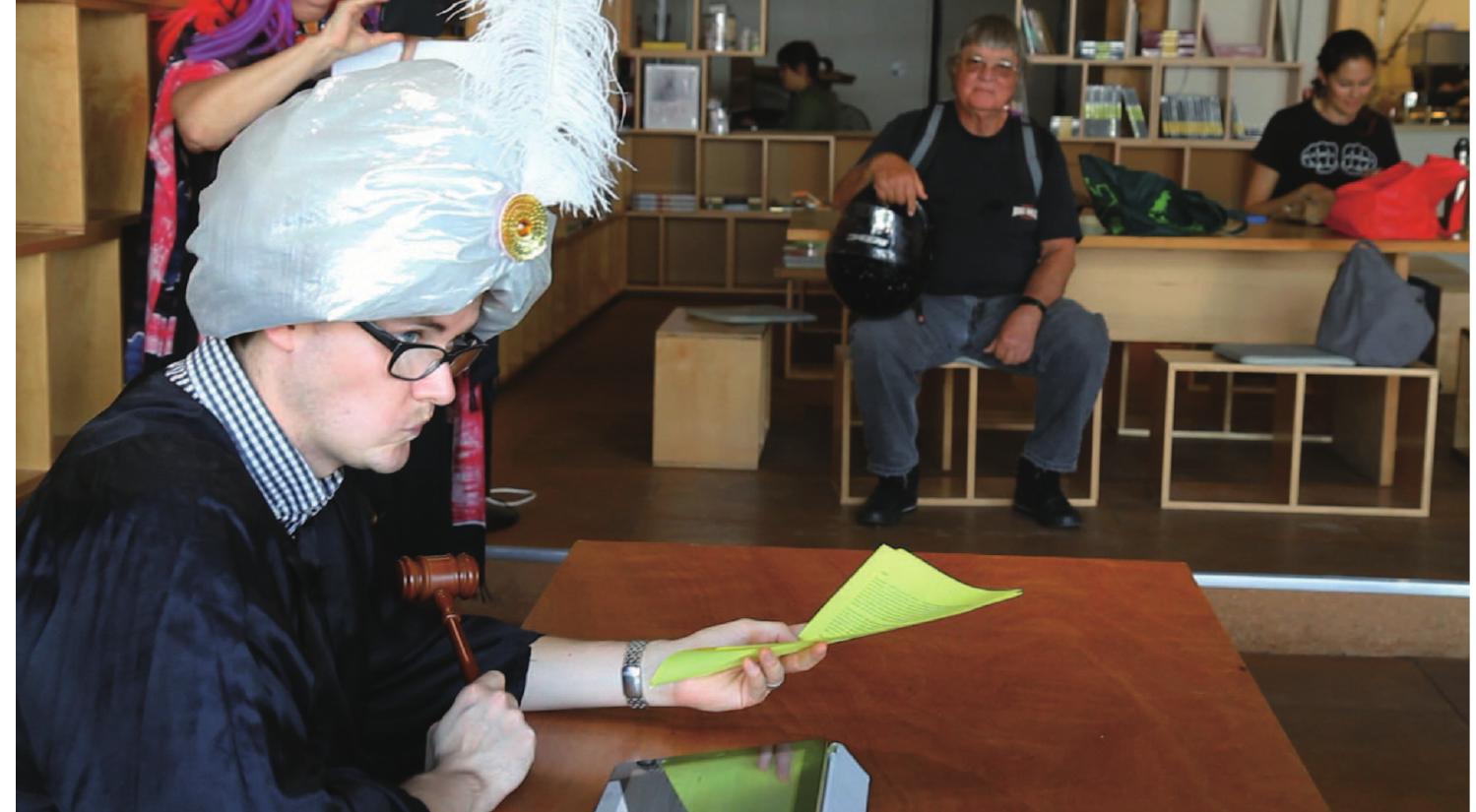 Figure 6. Costumed actor engaging Yellow/Green team as Dr. Dator looks on in the back ground  that in some scenarios “Yes” escalated and in others “No” escalated the conflict. In addition to having gamers vocalize their Yes/No answer, each gamer was also asked to provide a brief justification for their decision. While we thought it was important to recognize the systemic and institutional forces that shape power relations, we very much wanted to emphasize the power of everyday choices and the responsibility that comes along with access to varying degrees of communication technology. Both actors and gamers were furnished a complete script to follow along with during the scenario experience, and gamers were unaware of our escalate/deflate metric for recording decisions and were encouraged to inhabit their scenarios with abandon. 
