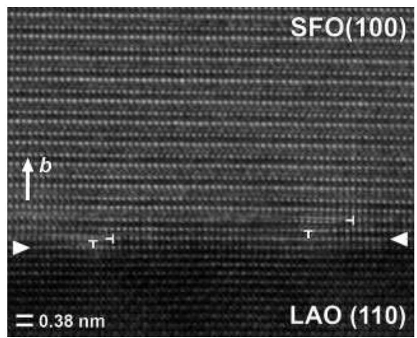 Figure 6. [100]seo HREM image of the SFO/LAO interface (indicated by white arrowheads) showing the presence of four dislocations associated to the presence of steps in the LAO substrate and the lattice mismatch between substrate and film. The average visible repeat period along the bspo-axis corresponds to the -SrO—FeOz-SrO—Fe202,5/2-SrO— sequence of alternating layers. 