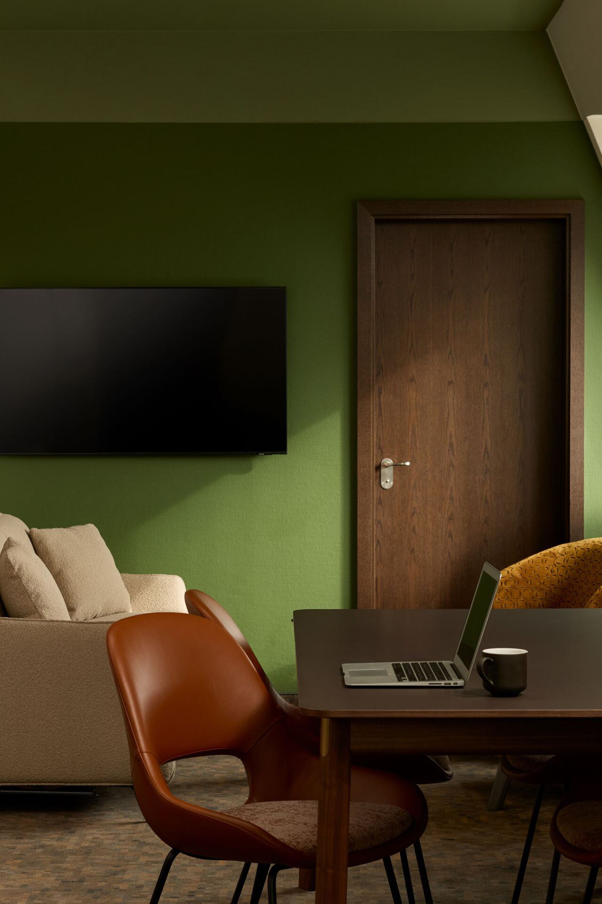 Hotel Frankfurt Metropolitan The Cloud One room with green painted wall, beige sofa, flat-screen TV, wooden table with laptop and coffee cup, as well as several chairs and a lamp.