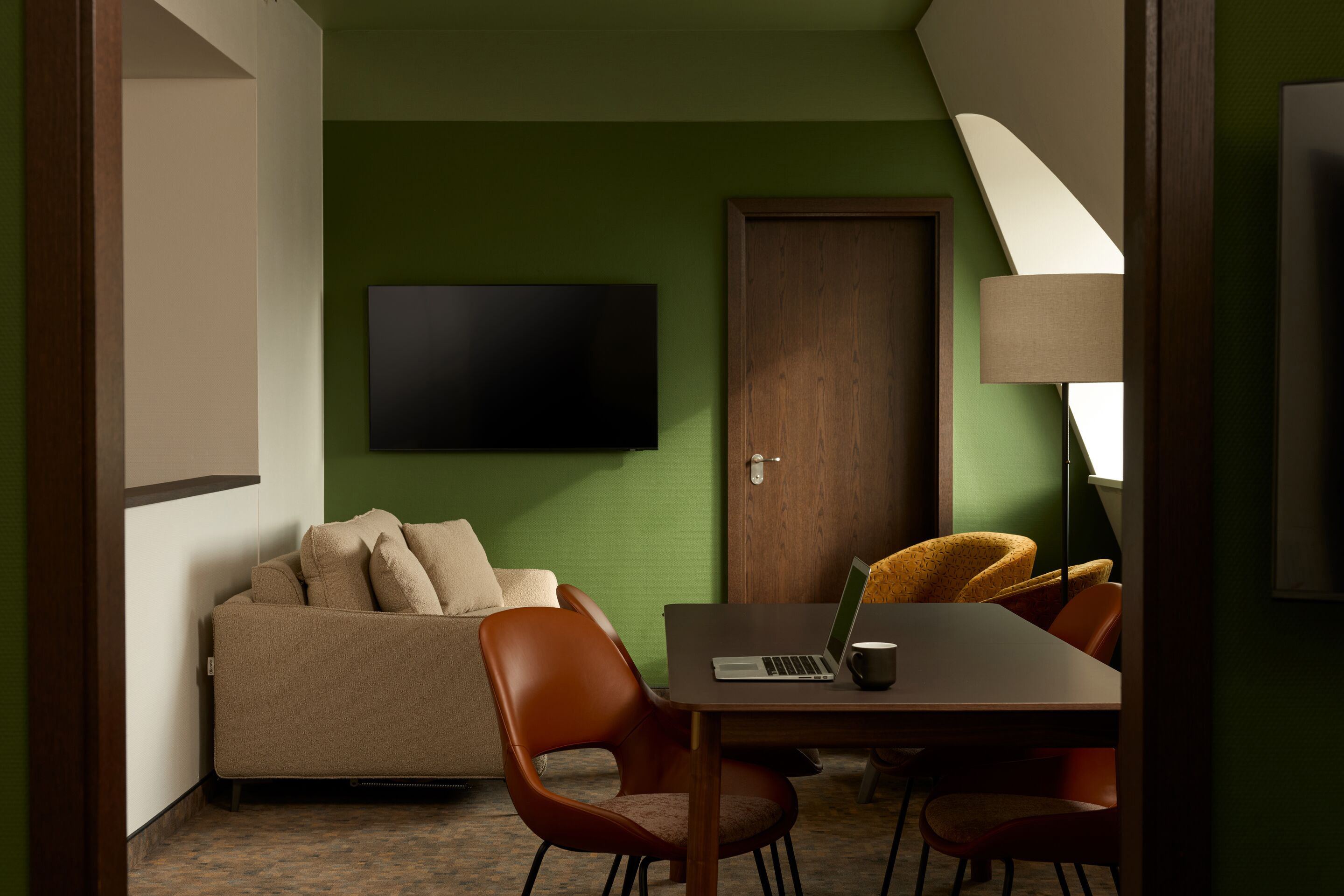 Hotel Frankfurt Metropolitan The Cloud One room with green painted wall, beige sofa, flat-screen TV, wooden table with laptop and coffee cup, as well as several chairs and a lamp. Hotel Frankfurt Metropolitan The Cloud One room with green painted wall, beige sofa, flat-screen TV, wooden table with laptop and coffee cup, as well as several chairs and a lamp.