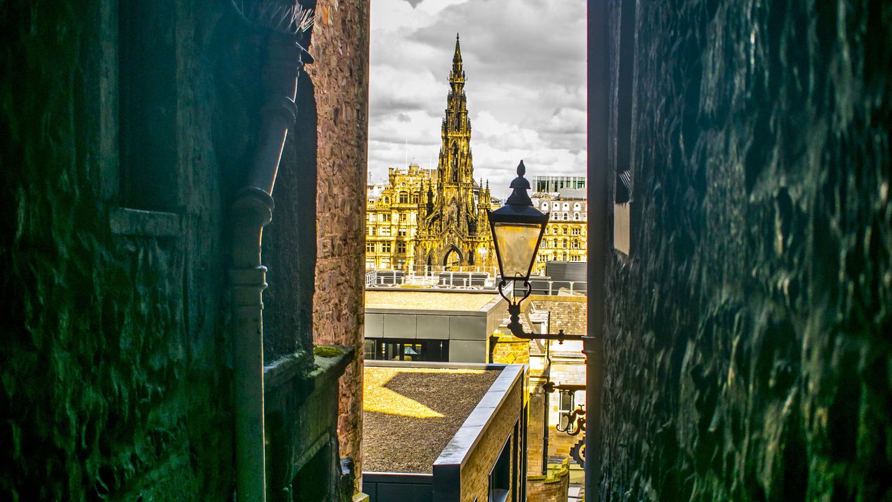 Blick durch einen engen, dunklen Durchgang auf eine historische Stadt mit einem hohen, gotischen Turm und einem Laternenpfahl bei bewölktem Himmel.
