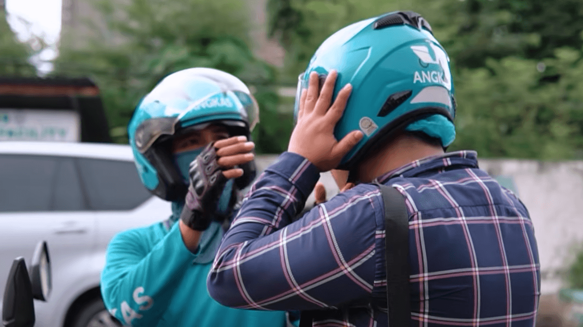An Angkas motorcycle driver in a teal uniform and helmet helps a passenger adjust their Angkas helmet