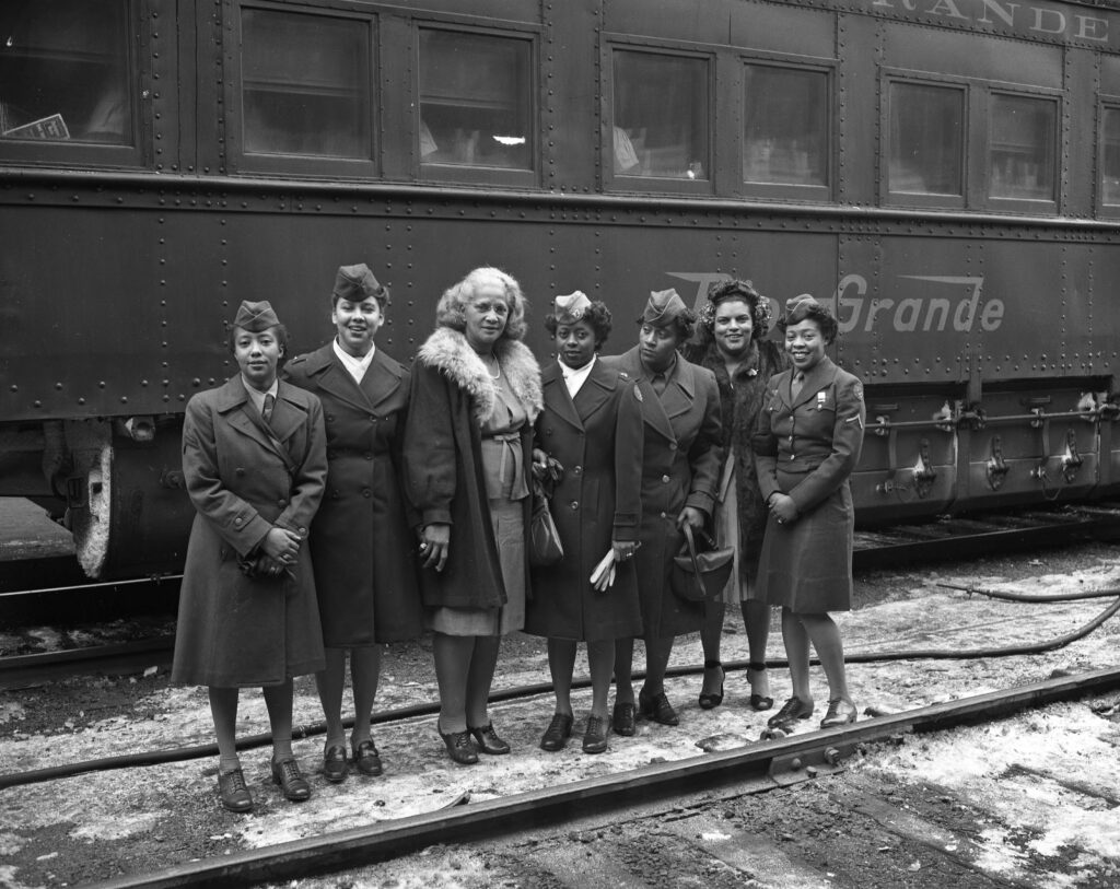 Seven women in military uniforms and a woman in a fur-collared coat stand smiling in front of a vintage train car labeled "Grande," conveying camaraderie.