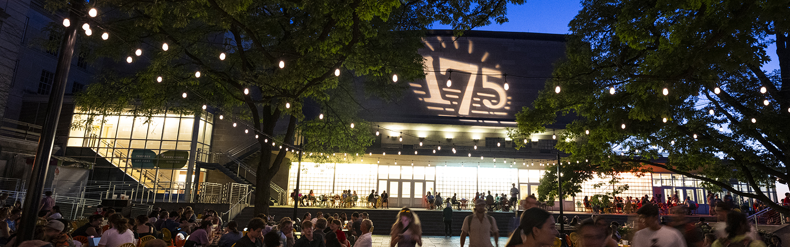 Evening view of people gathered on the Memorial Union Terrace, illuminated by string lights and the venue's illuminated facade featuring the UW 175 anniversary logo.