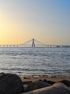 Sunset at Bandra-Worli Sea Link, with the bridge silhouetted against a warm sky and gentle waves along the rocky shore.
