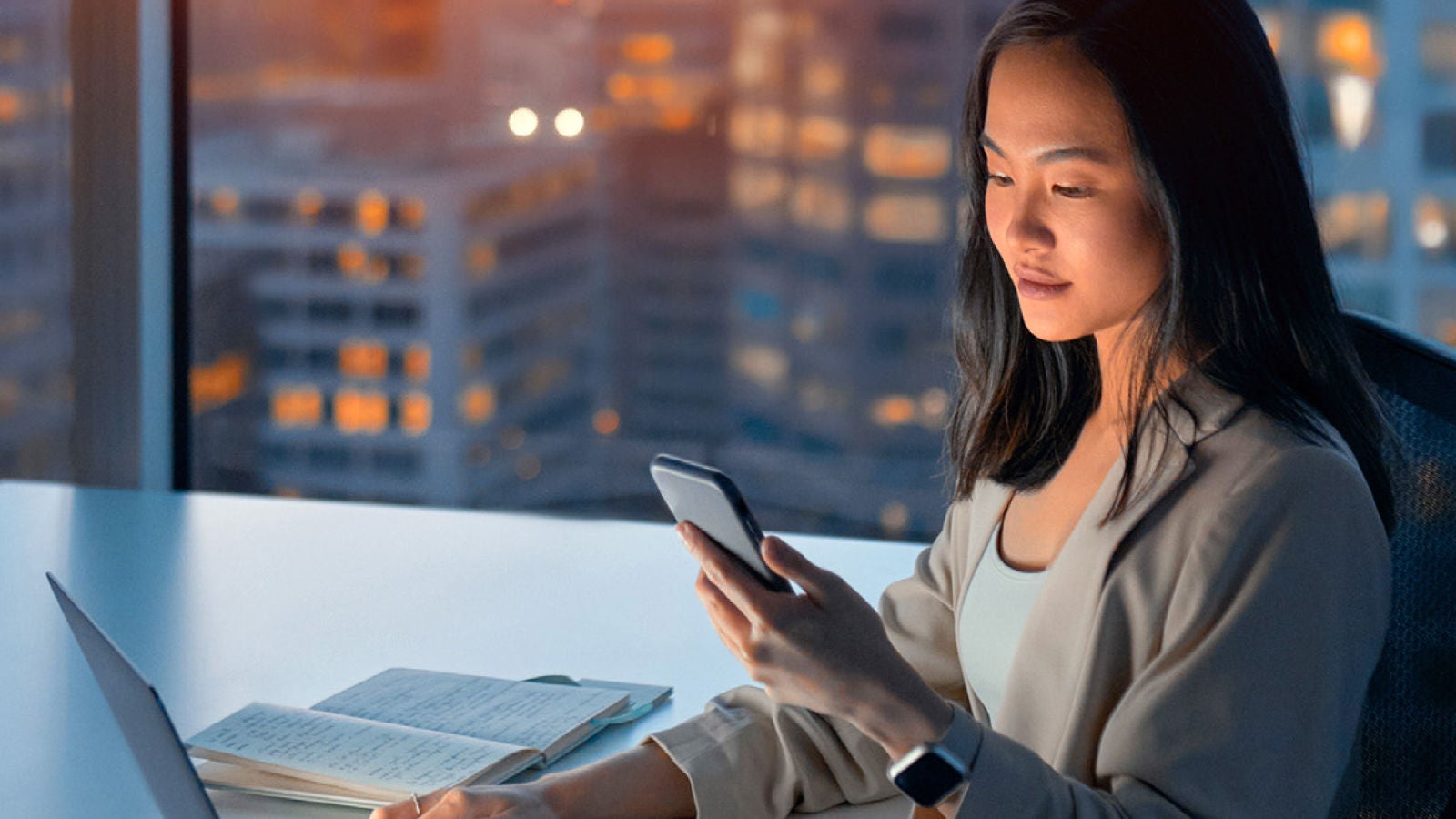 Woman working at a modern office desk in the evening. She is focused on a smartphone while using a laptop, with an open notebook beside her and city lights visible through the window.