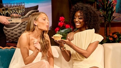 A woman blows out a candle on a cupcake being held by a second woman seated on a couch