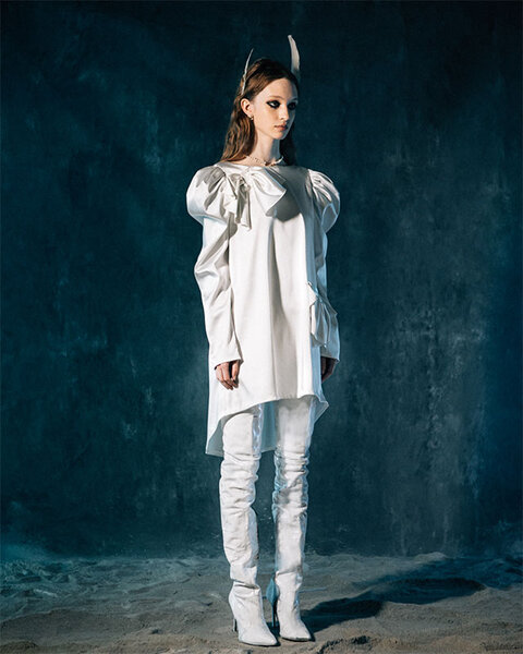 A model wears an all white ensemble with feathers in their hair while standing on a sea-blue backdrop.